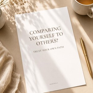 Puede incluir: Una impresión rectangular blanca con el texto gris oscuro COMPARING YOURSELF TO OTHERS? TRUST YOUR OWN PATH y la palabra MAISONELLA en la parte inferior. La impresión se muestra con un bolígrafo dorado, una taza de cerámica y tela beige.