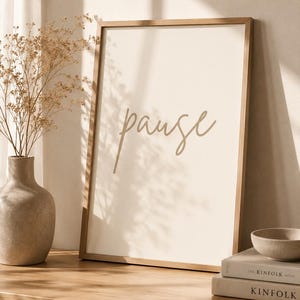 Puede incluir: Una impresión enmarcada con la palabra "pause" en una fuente cursiva. La impresión se muestra sobre una superficie de madera con un jarrón de flores secas y dos libros. La paleta de colores es neutra con tonos cálidos.