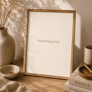 Puede incluir: Un marco rectangular de madera que contiene una impresión de color crema con el texto negro centrado "Keep going, gently". El marco está colocado en posición vertical, mostrando un diseño limpio con un borde fino, resaltando la tipografía.