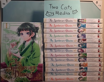 May include: A collection of The Apothecary Diaries manga volumes, including volume one as a standalone book and a stack of volumes two through fourteen. A light blue sign with the text Two Cats Media is positioned above the stacked books.