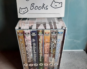 May include: A boxed set of seven A Silent Voice manga volumes by Kodansha Comics, arranged side-by-side. A white sign sits on top with the handwritten text Cat Cat Books and two cat face drawings. Each spine features a unique color and illustration.