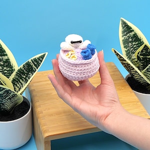 May include: A crocheted, light pink basket with a white character, yellow and blue accents. The basket is held in front of a blue background with two potted snake plants. The character has a black bar for eyes.