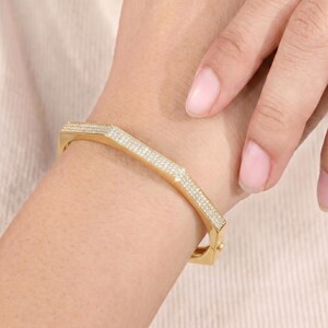 May include: A gold-toned hinged bangle bracelet featuring a geometric hexagonal shape. The outer surface of the bracelet is encrusted with numerous small, clear pav&eacute;-set stones that cover the entire visible top and side sections of the metal band.