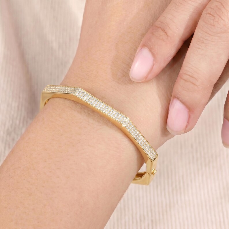 May include: A gold-toned hinged bangle bracelet featuring a geometric hexagonal shape. The outer surface of the bracelet is encrusted with numerous small, clear pav&eacute;-set stones that cover the entire visible top and side sections of the metal band.