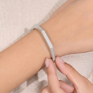 May include: A silver-toned hinged bangle bracelet featuring a geometric hexagonal shape. The entire outer surface of the rigid metal band is encrusted with numerous small, clear, sparkling stones that catch the light, creating a uniform, brilliant finish.