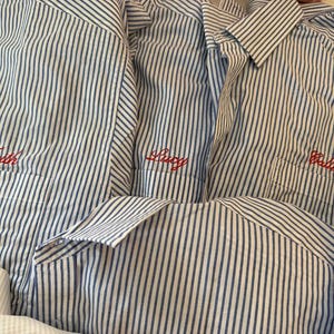Peut inclure: Plusieurs chemises à rayures bleues et blanches avec des noms brodés en rouge sur les poches. Les noms "Faith", "Lucy" et "Callie" sont visibles. Les chemises ont des cols boutonnés et des manches longues.