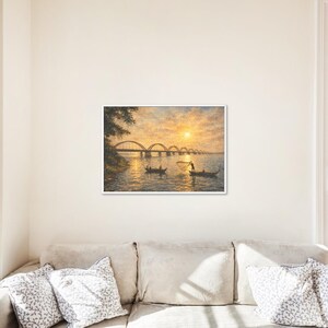 May include: A framed landscape painting depicts a river scene at sunset. The artwork features a bridge with multiple arches, boats with people, and a vibrant sky with a bright sun. The painting is displayed above a beige sofa.