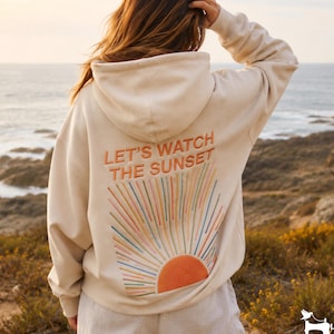 Può includere: Una felpa con cappuccio color crema caratterizzata da un ricamo sul retro. Il testo "LET'S WATCH THE SUNSET" è posizionato sopra un grafico solare stilizzato con linee radianti nei toni dell'arancione, rosa, blu e giallo.