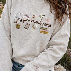 Op de afbeelding: Een beige sweater met ronde hals en bruin geborduurde tekst let a girl read in peace. Het ontwerp bevat geborduurde iconen van een koptelefoon, bril, mok, strik, sokken, kaars, e-reader, bloemen en boeken.