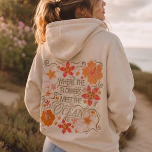 May include: A cream-colored hooded sweatshirt featuring an embroidered design on the back. The embroidery includes orange, red, and white tropical flowers, seashells, and the text "WHERE THE FLOWERS MEET THE OCEAN" in a decorative font.