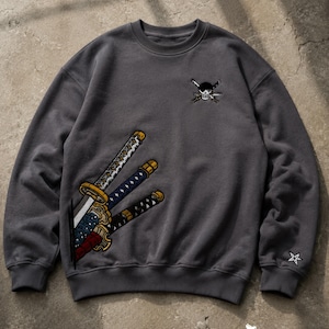 Könnte beinhalten: Ein anthrazitfarbenes Sweatshirt mit Rundhalsausschnitt, das auf der rechten Brust ein gesticktes Totenkopf-Emblem und unten links eine große Grafik mit drei Katana-Schwertern zeigt. Am linken Ärmelbündchen befindet sich ein kleines Sternsymbol.