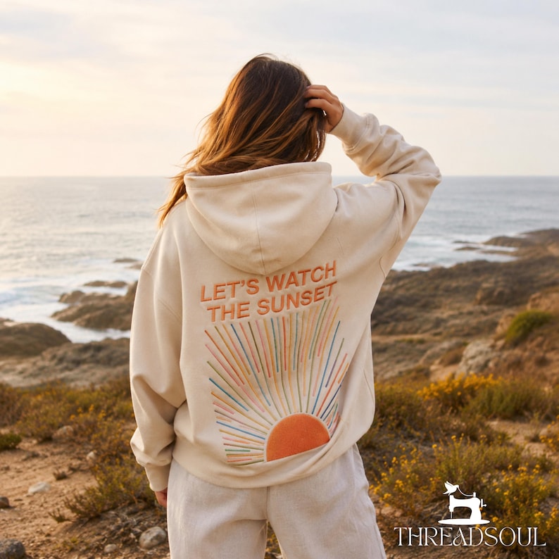 May include: A cream-colored hooded sweatshirt featuring an embroidered design on the back. The text reads "LET'S WATCH THE SUNSET" above a stylized sun graphic with radiating lines in shades of orange, pink, blue, and yellow.