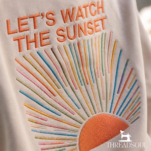 May include: A beige sweatshirt featuring embroidered text that reads LET'S WATCH THE SUNSET in orange. Below the text is a semi-circular orange sun with radiating lines in various colors including blue, green, pink, and yellow, creating a sunburst design.