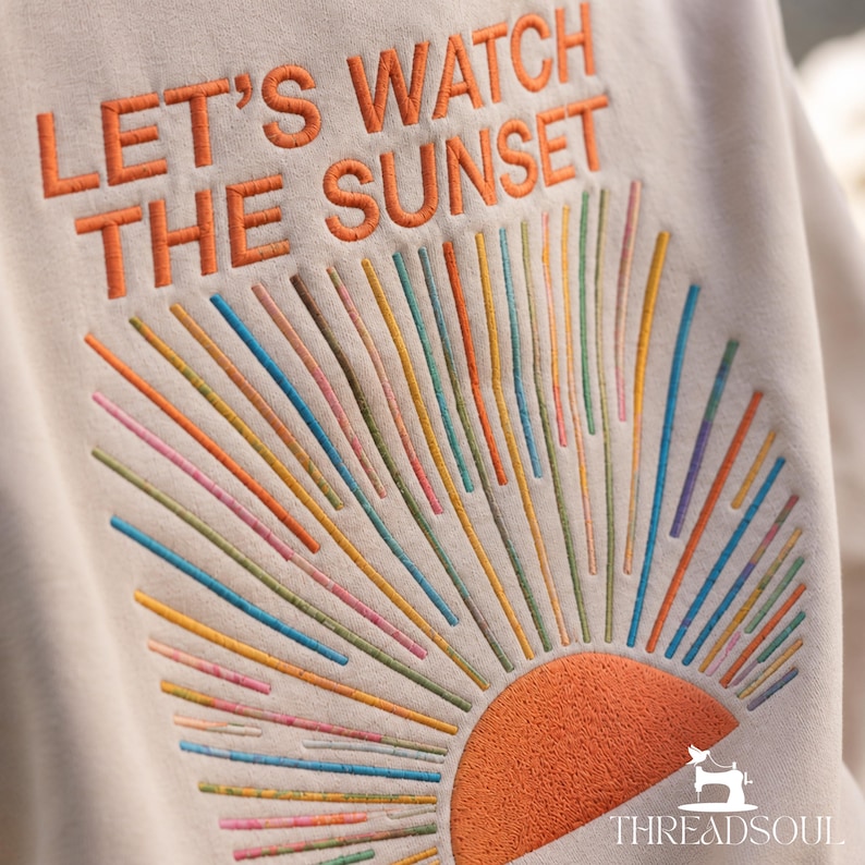 May include: A beige sweatshirt featuring embroidered text that reads LET'S WATCH THE SUNSET in orange. Below the text is a semi-circular orange sun with radiating lines in various colors including blue, green, pink, and yellow, creating a sunburst design.