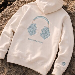 May include: An off-white hooded sweatshirt featuring an embroidered design on the back of light blue headphones with seashell ear cups. Below the headphones, the text reads "listen to the ocean" in blue. A small seashell is embroidered on the left sleeve.