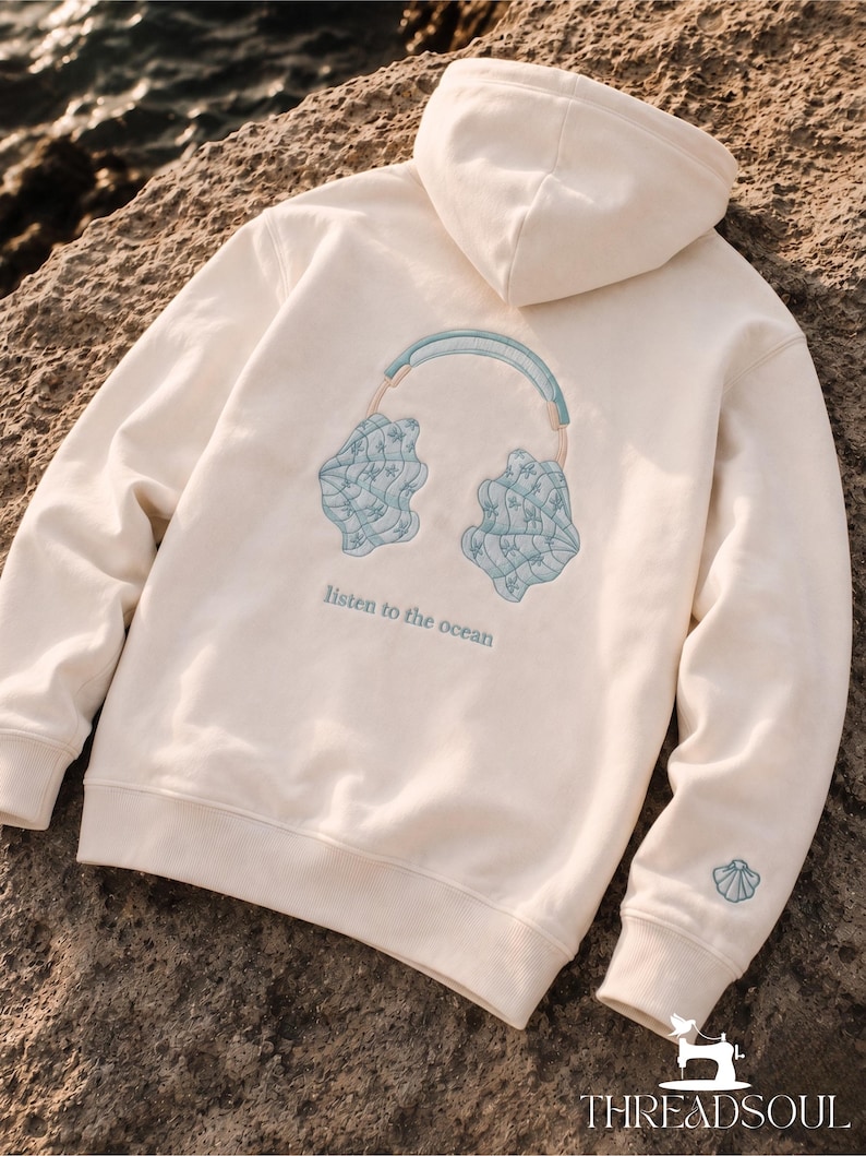 May include: An off-white hooded sweatshirt featuring an embroidered design on the back of light blue headphones with seashell ear cups. Below the headphones, the text reads "listen to the ocean" in blue. A small seashell is embroidered on the left sleeve.