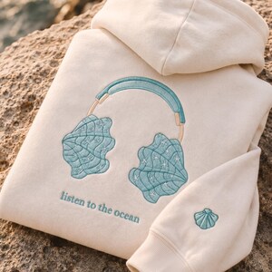 May include: A cream-colored hooded sweatshirt featuring an embroidered design of teal seashell headphones. Below the headphones, the text reads "listen to the ocean" in teal. A small teal seashell is embroidered on the sleeve cuff.