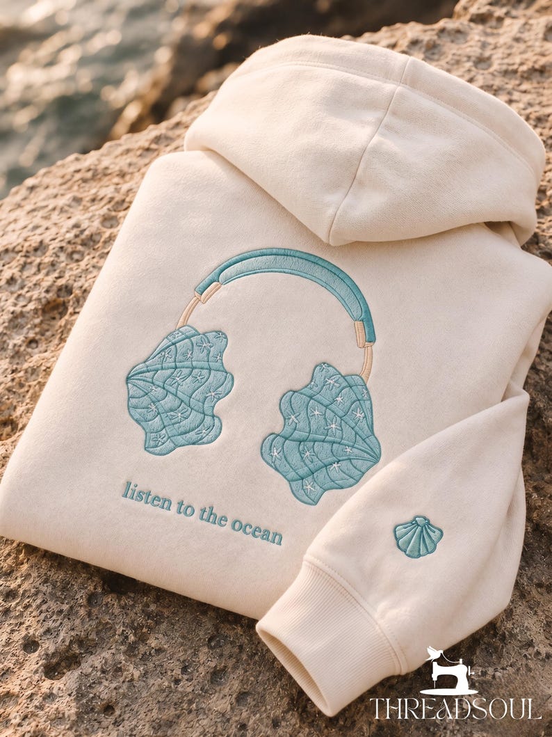 May include: A cream-colored hooded sweatshirt featuring an embroidered design of teal seashell headphones. Below the headphones, the text reads "listen to the ocean" in teal. A small teal seashell is embroidered on the sleeve cuff.
