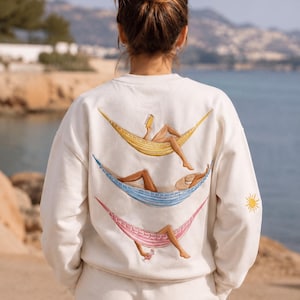 May include: An off-white crewneck sweatshirt featuring an embroidered design on the back of three colorful hammocks, each occupied by a figure. A yellow sun is embroidered on the sleeve. The hammocks are yellow, blue, and pink with detailed figures.