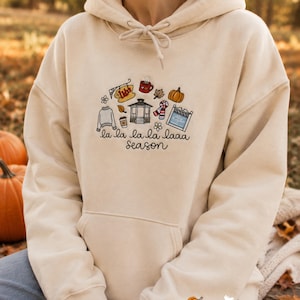 Puede incluir: Una sudadera con capucha de color crema que presenta un gráfico bordado central de un cenador, una taza de café, una calabaza y un letrero que dice Luke's, con el texto la la la la laaa season debajo. Una pequeña calabaza naranja está bordada en la manga.