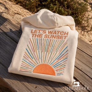 May include: A cream-colored hooded sweatshirt featuring an embroidered design on the back. The text "LET'S WATCH THE SUNSET" is stitched in orange above a sun graphic with rays in various colors including blue, green, and orange.