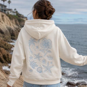 May include: An off-white hooded sweatshirt featuring a light blue graphic print on the back. The design includes a palm tree, a large hibiscus flower, various seashells, and two starfish. The hoodie has long sleeves and a ribbed hem and cuffs.