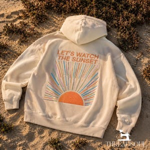 May include: An off-white hooded sweatshirt featuring an embroidered design on the back. The text reads "LET'S WATCH THE SUNSET" above a semi-circular orange sun with radiating multi-colored embroidered lines in blue, pink, and yellow hues.