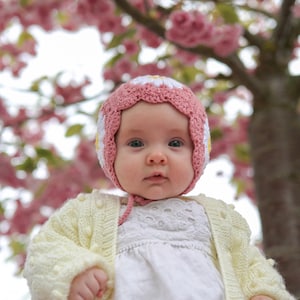 Peut inclure: Un bébé portant un bonnet rose et blanc avec un motif de marguerite et un cardigan jaune clair. Le bébé porte une robe blanche. Le mot "Wollie" est en haut de l'image. L'arrière-plan est un arbre floral rose flou.