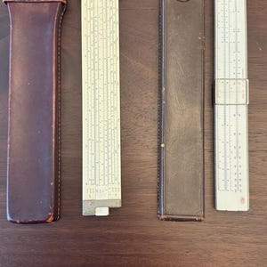 Puede incluir: Cuatro reglas de cálculo antiguas y sus estuches se muestran sobre una superficie de madera. Las reglas de cálculo tienen escalas y marcas visibles. Los estuches son de cuero y de diferentes tonos marrones.