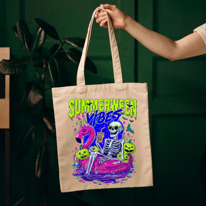 以下が含まれることがあります： ベージュのトートバッグ。カラフルなグラフィックデザインで、「SUMMERWEEN VIBES」の文字、サングラスをかけた骸骨がピンクのフラミンゴの浮き輪に乗っている様子、カボチャ、コウモリが描かれています。