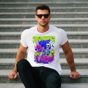 以下が含まれることがあります： 白いTシャツ。カラフルなグラフィックで、フラミンゴの浮き輪に乗った骸骨、カボチャ、そして「Summerween Vibes」の文字が描かれています。デザインは緑、青、ピンク、紫のアクセントがあります。