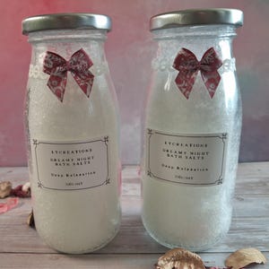 Puede incluir: Dos botellas de vidrio llenas de sales de baño blancas, cada una con una tapa plateada y un lazo rojo decorativo. Las botellas tienen etiquetas que dicen "Dreamy Night Bath Salts" y "Deep Relaxation". Las botellas son de 200g netos.