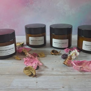 May include: Four brown glass jars with black lids, labelled "Breath Easy Chest Rub." The jars are on a light wood surface, with dried flowers and petals. Each jar contains 28g of product.