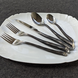 May include: A white, scalloped-edge serving platter holds a set of silverware. The set includes two forks, a knife, and two spoons. The silverware has dark, textured handles and shiny, silver heads.