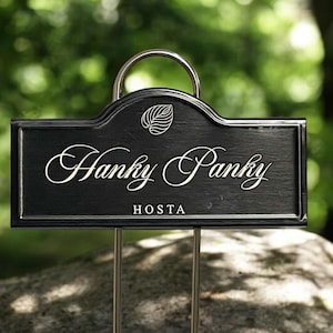 May include: Black metal garden marker with a silver metal loop and a white leaf design. The marker has the text "Hanky Panky HOSTA" written on it.
