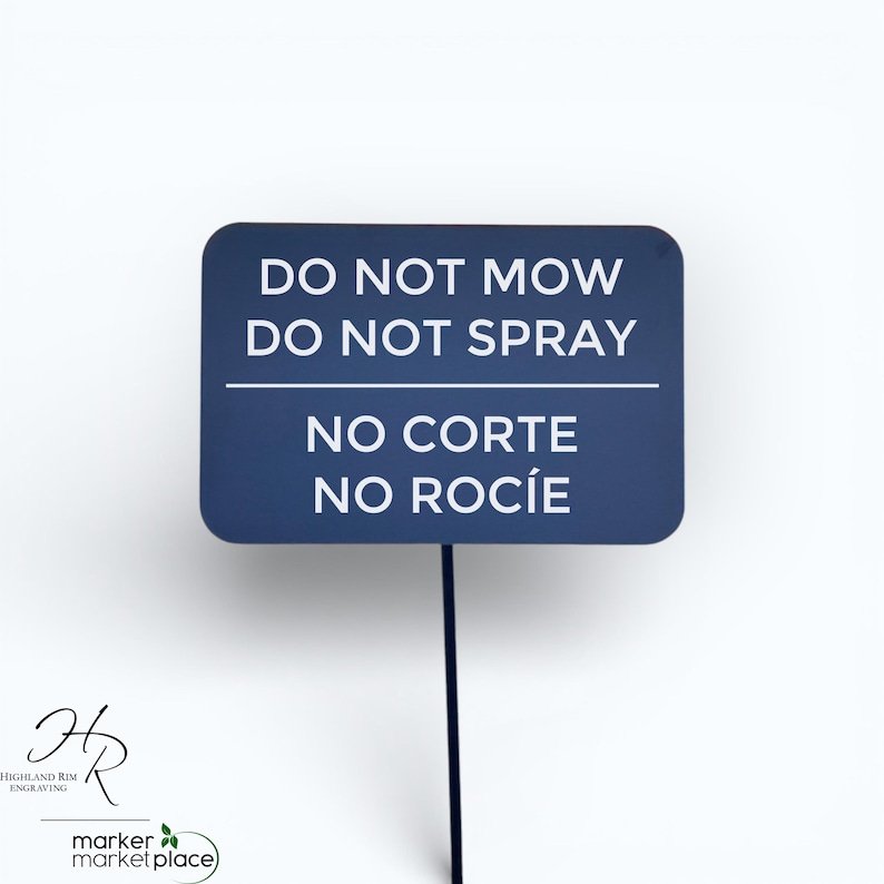 May include: A dark blue rectangular sign on a black stake. The sign reads "DO NOT MOW DO NOT SPRAY" in white text, with "NO CORTE NO ROC&Iacute;E" below. The sign is for a garden or lawn.
