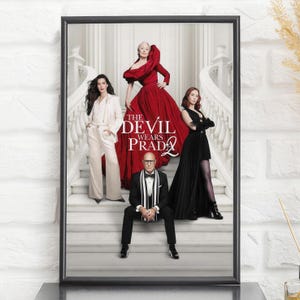 May include: A framed movie poster for The Devil Wears Prada 2 featuring four individuals on a white staircase. One person wears a red gown, one a white suit, one a black dress with gloves, and one a black tuxedo with a striped scarf.
