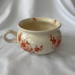 May include: Cream-coloured ceramic bowl with a handle, decorated with a floral pattern in shades of red and pink. The bowl has a rounded shape and a slightly flared rim. A classic piece for serving or display.
