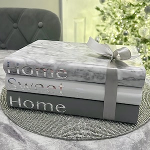 May include: A stack of decorative books with the words "Home Sweet Home" in silver lettering. The top book has a marble effect, and the set is tied with a silver ribbon. The books are on a beaded placemat.