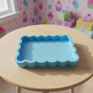 Op de afbeelding: Een lichtblauw rechthoekig dienblad met een geschulpte rand staat op een lichtbruine houten tafel. De lade is leeg en heeft een glad oppervlak. De achtergrond heeft een roze behang met cupcake- en ballonontwerpen.