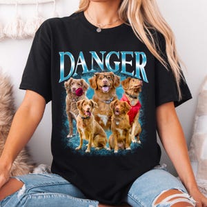 May include: A black short-sleeved t-shirt featuring a graphic print of five brown dogs. The word DANGER is printed in large blue metallic-style letters above the dogs. One dog wears glasses, one holds a red rose, and one wears a red vest.