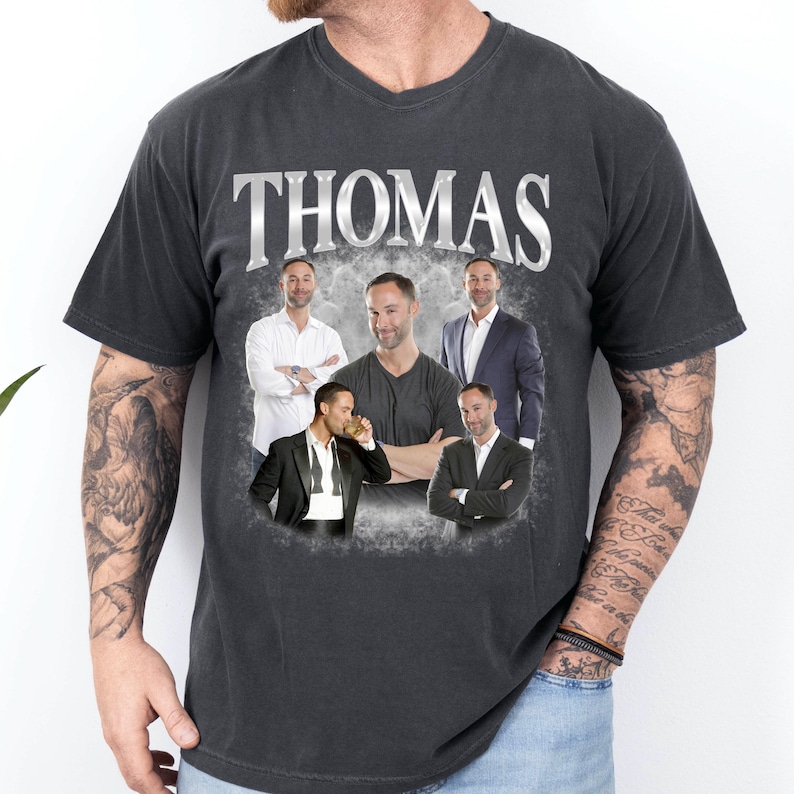 May include: A charcoal gray short-sleeve t-shirt featuring the name THOMAS in large silver lettering at the top. Below the text, a collage of five photographs shows the same individual in various outfits, including a white shirt and suits.