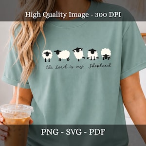 Può includere: Una t-shirt verde salvia con una grafica di cinque pecore bianche con facce e zampe nere in varie pose. Sotto le pecore, il testo nero in corsivo recita, the Lord is my Shepherd. La maglietta ha maniche corte e un girocollo.