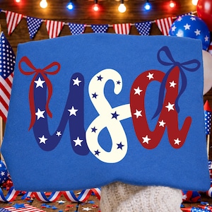 May include: A blue textile item with the word "USA" in white, red, and blue, decorated with stars and bows. The letters are outlined with red and blue ribbons and white stars. The background includes American flags and festive decorations.