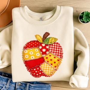 May include: Cream-colored sweatshirt featuring a colorful apple design. The apple is made of patchwork fabric with red, yellow, and white patterns, including hearts, daisies, and gingham. The sweatshirt has a crew neck and long sleeves.