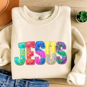Puede incluir: Sudadera color crema con la palabra "JESUS" en letras coloridas estilo patchwork. Las letras presentan varios patrones, incluyendo florales, cuadros y lunares. La sudadera está doblada sobre una superficie de madera.