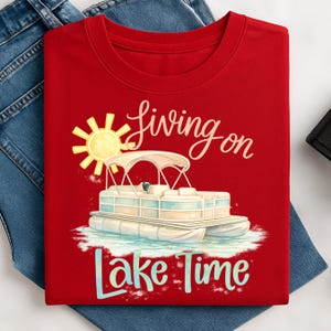 May include: A red t-shirt featuring a graphic of a pontoon boat on water with a sun illustration. The text reads "Living on Lake Time" in a script font. The shirt is displayed on a pair of blue jeans.