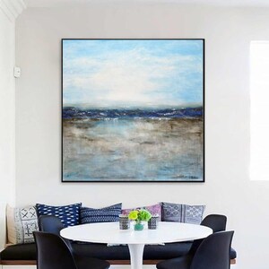May include: Abstract painting of a seascape with blue and brown hues. The painting depicts a horizon with a blue sky and a body of water with a sandy shore. The painting is framed in black and hangs on a white wall above a dining table.