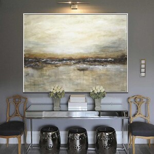 May include: A large abstract painting with a landscape theme. The painting features a muted color palette of browns, grays, and whites. The painting is hanging on a gray wall above a silver console table with three black stools.