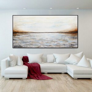 May include: A white sectional sofa with a red throw blanket in front of a large abstract painting of a seascape in shades of brown, white, and blue. The painting is hanging on a white wall in a modern living room.